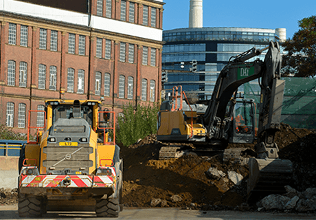 Yellow loading shovel and excavator next to a pile or rubble with buildings in the background