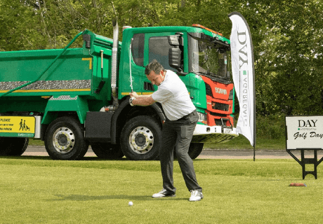 man swinging a golf club, with a green Day aggregates lorry in background