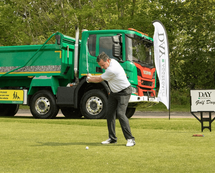 man swinging a golf club, with a green Day aggregates lorry in background